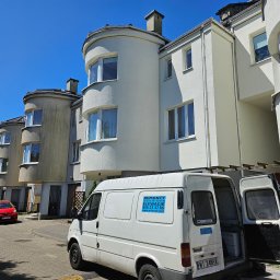 Miwobud - Ulica z szeregiem domów o charakterystycznych zaokrąglonych elementach architektonicznych, biały bus z otwartymi drzwiami i naklejką reklamującą usługi remontowo-elewacyjne, czerwony samochód w tle.