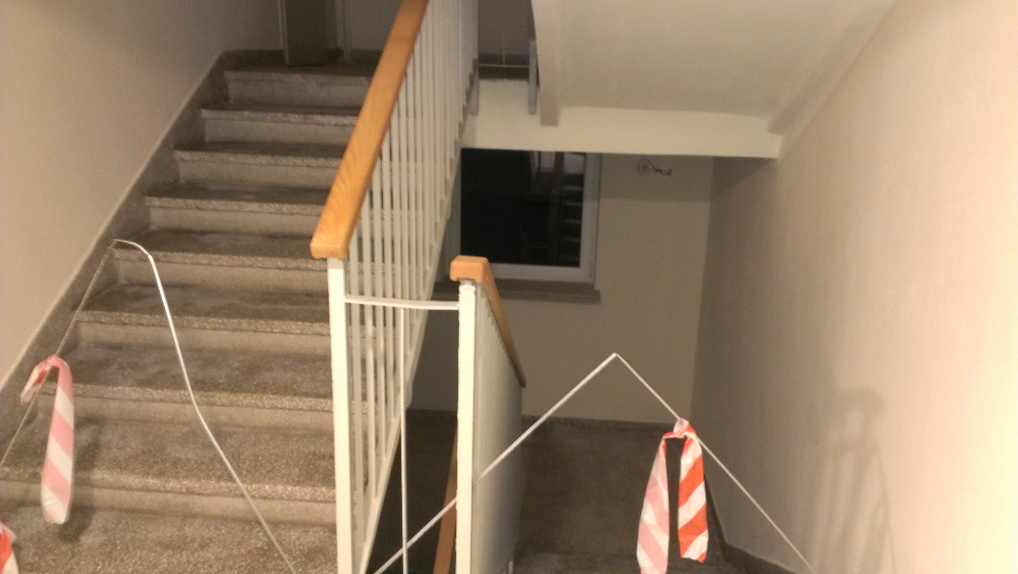 Klatka schodowa z lastryko, zabezpieczona biało-czerwoną taśmą, białe balustrady z drewnianymi poręczami, widok z góry.