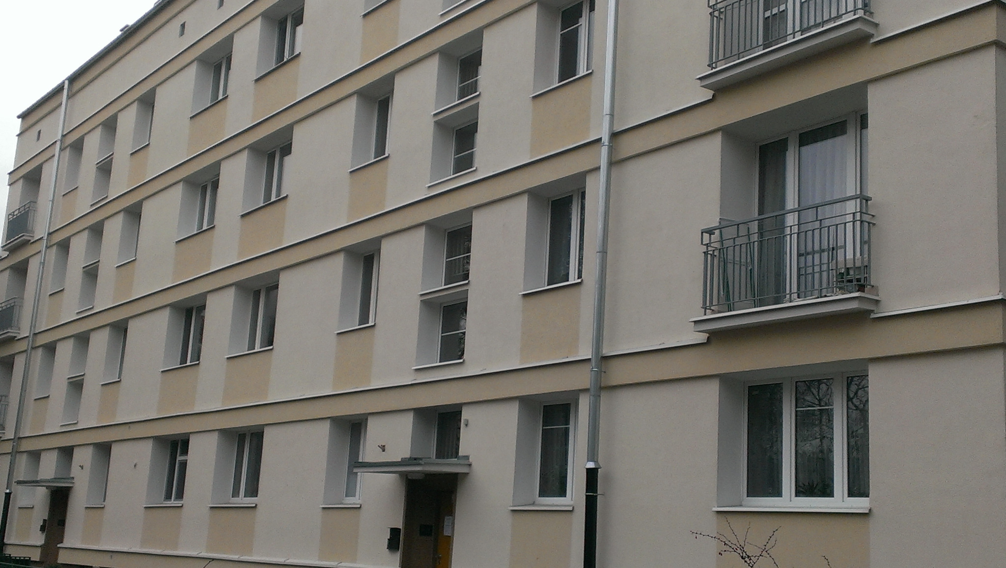 Odnowiona elewacja budynku wielorodzinnego z jasnobeżowymi pasami, widoczne balkony z metalowymi barierkami i rury spustowe.