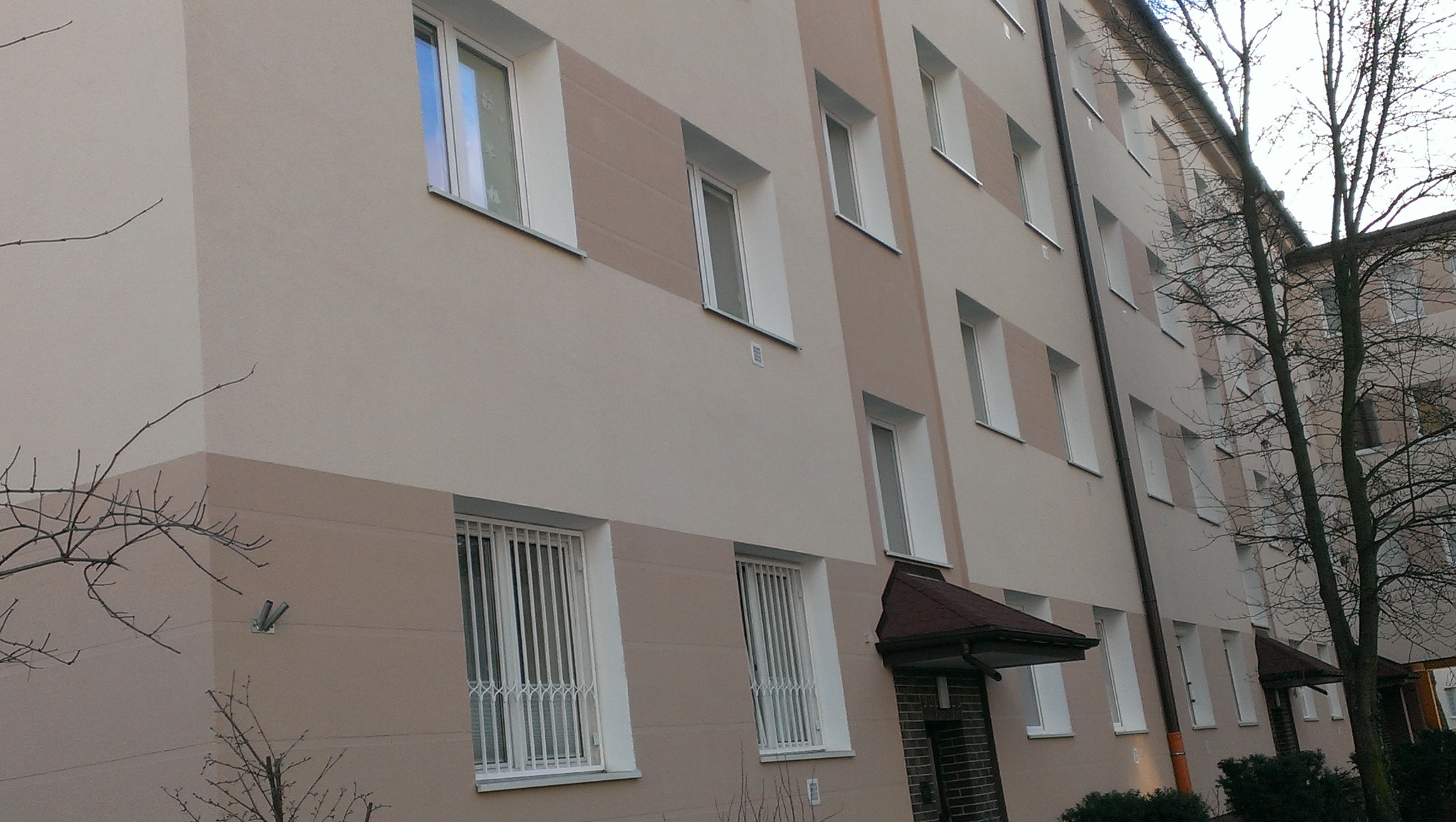 Wielopiętrowy budynek z elewacją w odcieniach beżu, z białymi ramami okiennymi i kratami w oknach na parterze, widoczny z perspektywy lekko od dołu.