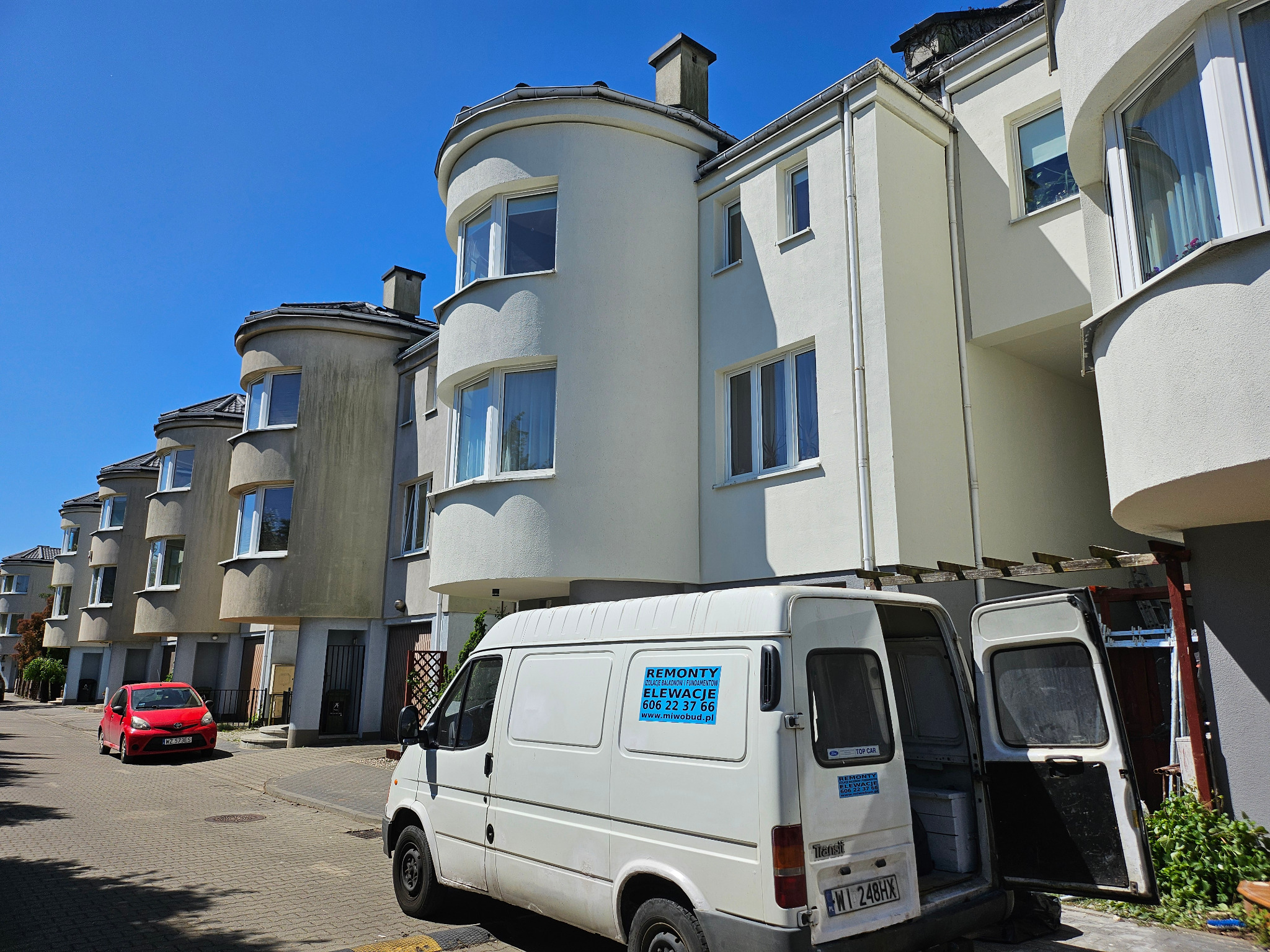 Ulica z szeregiem domów o charakterystycznych zaokrąglonych elementach architektonicznych, biały bus z otwartymi drzwiami i naklejką reklamującą usługi remontowo-elewacyjne, czerwony samochód w tle.