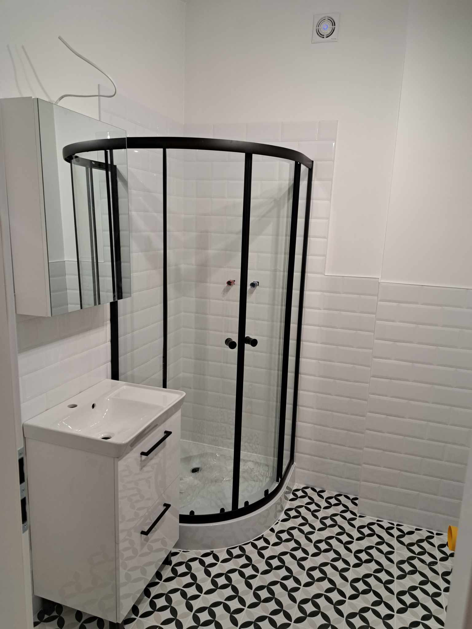 Nowoczesna łazienka z kabiną prysznicową w czarnej ramie, białą umywalką z szafką i wzorzystą podłogą w odcieniach szarości i czerni. Białe płytki imitujące cegiełki na ścianach.