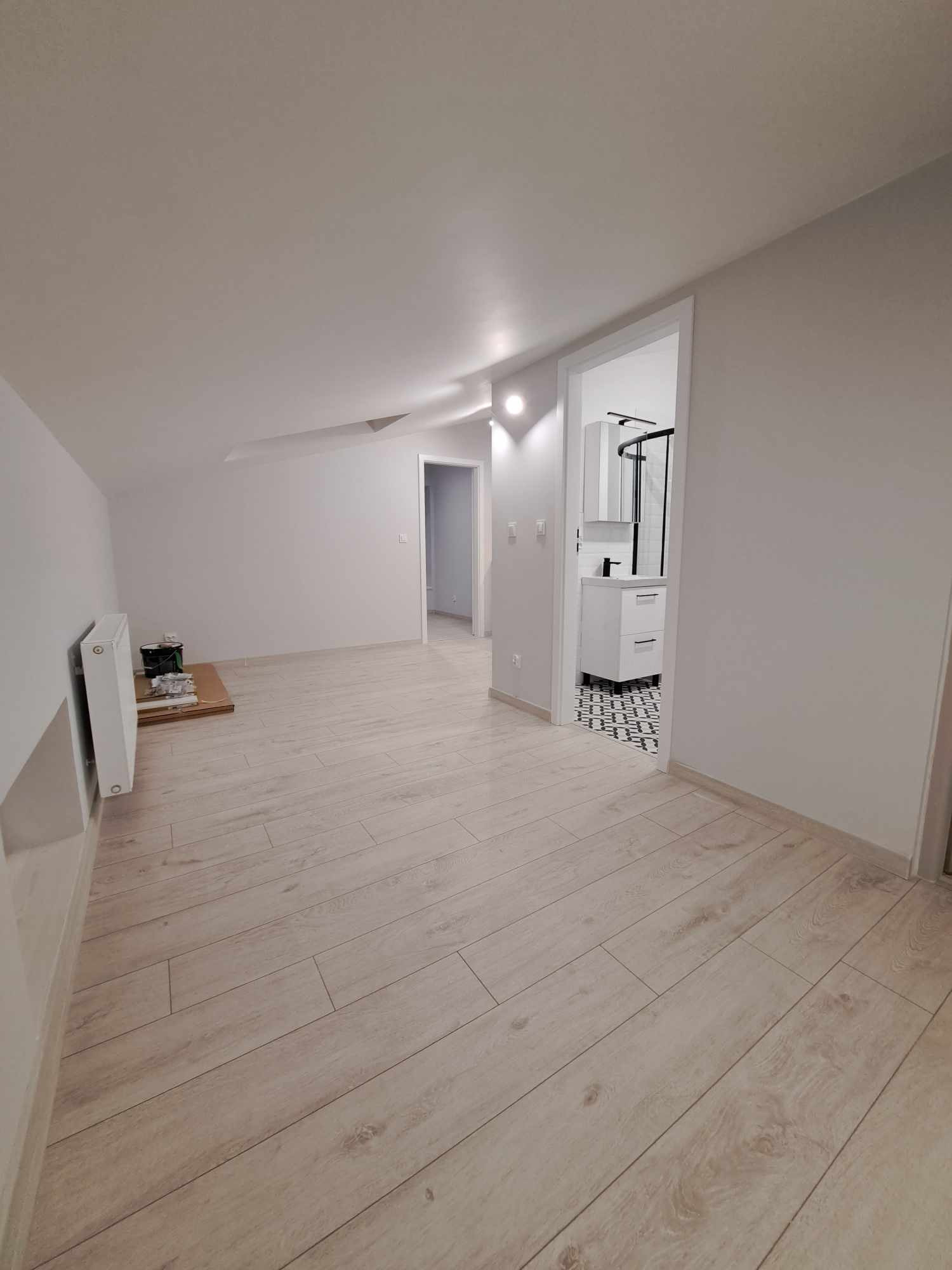 Jasne panele podłogowe w odnowionym pokoju na poddaszu z widokiem na łazienkę z kabiną prysznicową typu walk-in i białą szafką umywalkową. Na podłodze w łazience czarno-biała mozaika.