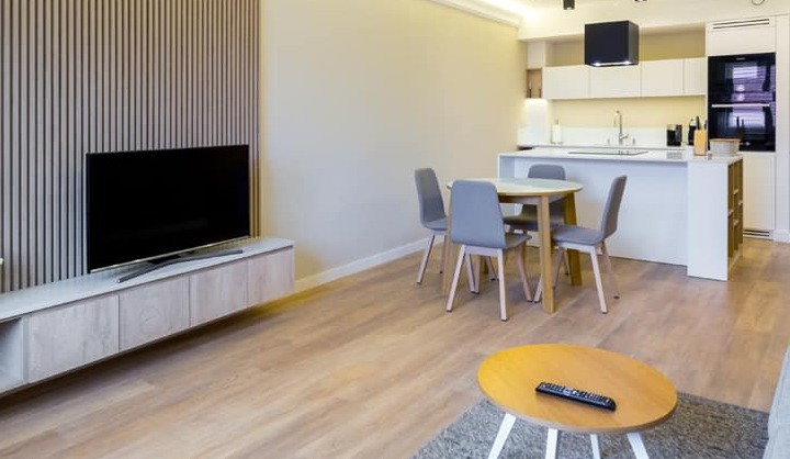 Jasne, nowocześnie urządzone wnętrze mieszkania z drewnianą podłogą, panelem ściennym za telewizorem, okrągłym stołem jadalnym z szarymi krzesłami i aneksem kuchennym w tle.