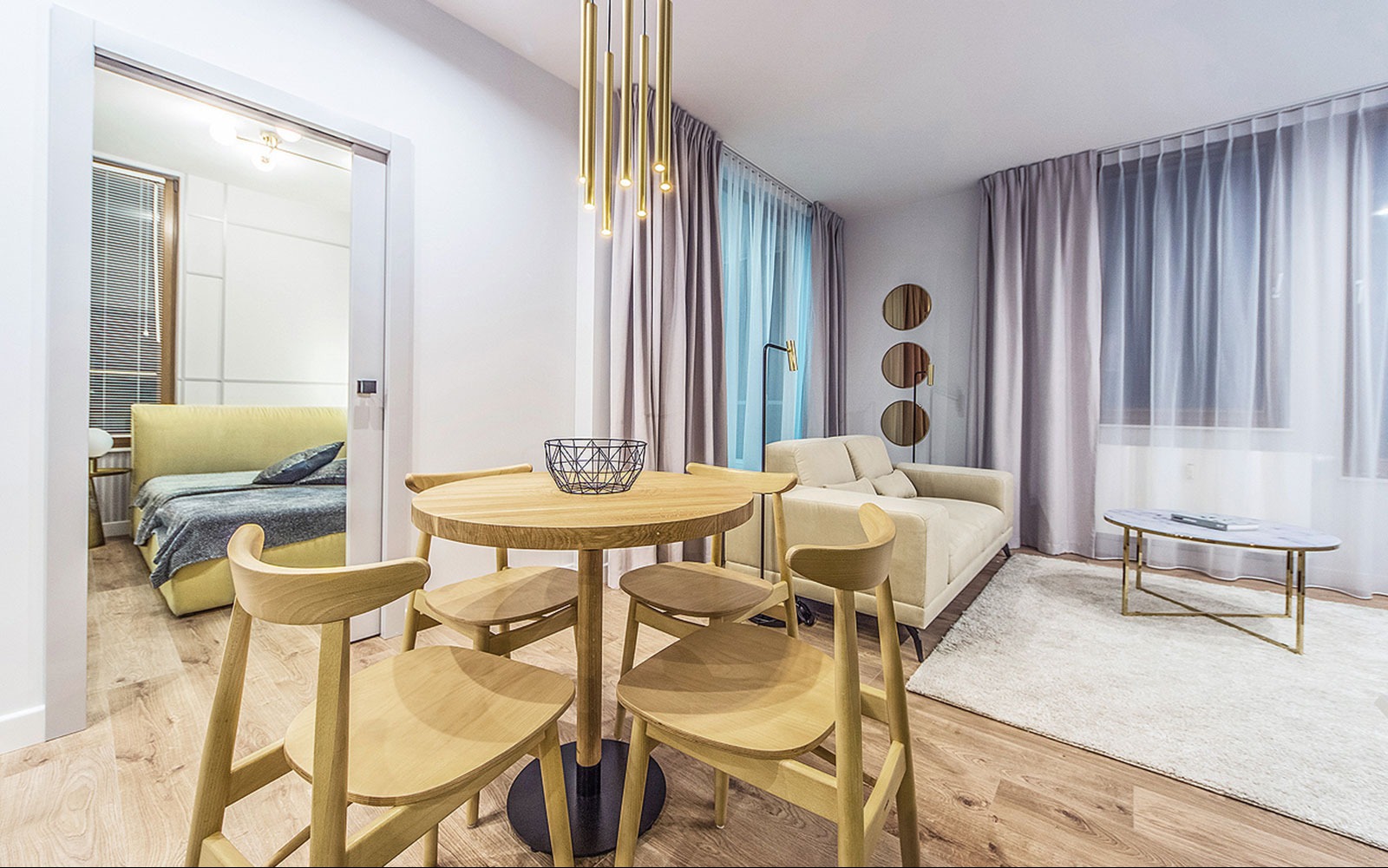Jasne, nowocześnie urządzone wnętrze mieszkania z okrągłym drewnianym stołem, krzesłami z jasnego drewna, beżową sofą i widokiem na sypialnię przez otwarte drzwi.