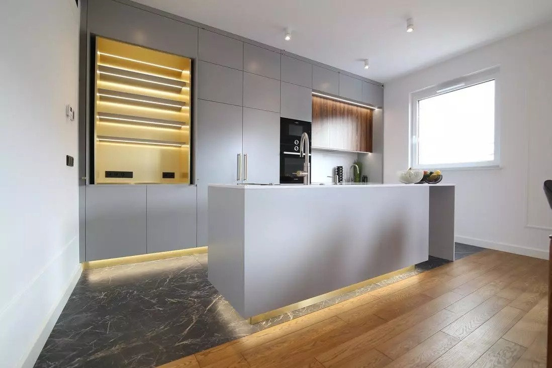 Nowoczesna kuchnia z szarymi szafkami, wyspą kuchenną z oświetleniem LED i marmurową podłogą przechodzącą w drewniany parkiet.