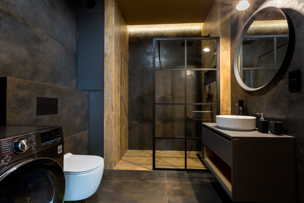 Nowoczesna łazienka w ciemnych odcieniach z kabiną prysznicową typu walk-in z czarnymi szprosami, okrągłym lustrem z podświetleniem LED, umywalką nablatową i pralką LG w rogu.