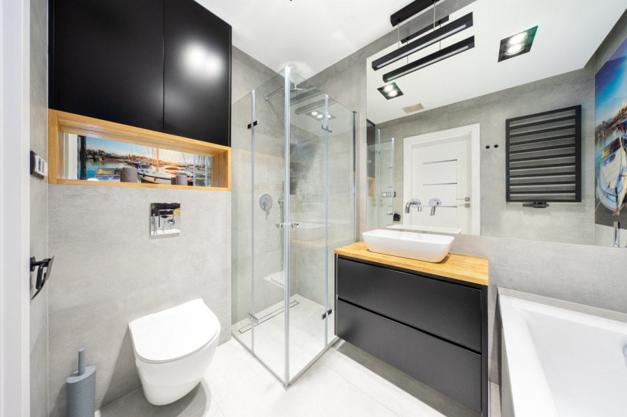 Nowoczesna łazienka z kabiną prysznicową, umywalką nablatową na drewnianym blacie i podwieszaną toaletą, szare płytki, czarne szafki i dekoracyjny panel z motywem portu.