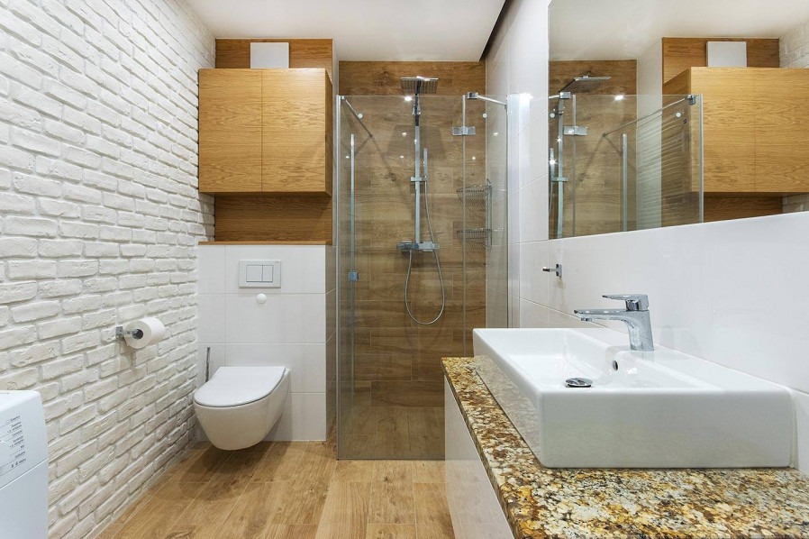Nowoczesna łazienka z kabiną prysznicową typu walk-in, umywalką nablatową na blacie z granitu, toaletą podwieszaną i ścianą z białej cegły dekoracyjnej.
