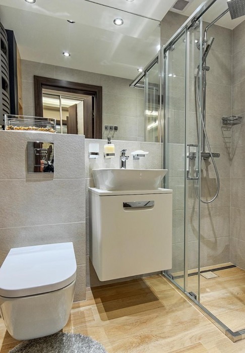 Nowoczesna łazienka z wiszącą toaletą, umywalką nablatową na szafce i kabiną prysznicową z przesuwnymi drzwiami, wykończona płytkami w odcieniach szarości i drewnopodobną podłogą.