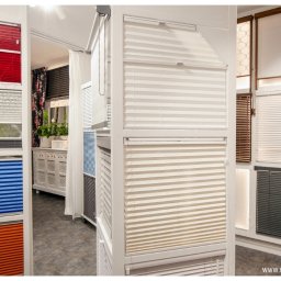 ROLETY AKCES - Ekspozycja różnokolorowych i różnorodnych typów osłon okiennych w salonie, widoczne plisy w odcieniach czerwieni, zieleni, niebieskiego i pomarańczowego, oraz żaluzje w kolorach białym, beżowym...