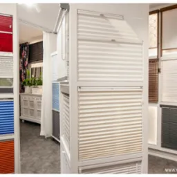 Ekspozycja różnokolorowych i różnorodnych typów osłon okiennych w salonie, widoczne plisy w odcieniach czerwieni, zieleni, niebieskiego i pomarańczowego, oraz żaluzje w kolorach białym, beżowym...