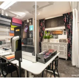 Wnętrze salonu z ekspozycją różnych typów przesłon okiennych, w tym żaluzji drewnianych i aluminiowych, z paletą kolorów na blacie i transparentną przegrodą ochronną.