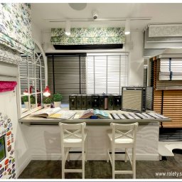 ROLETY AKCES - Ekspozycja różnych typów osłon okiennych w salonie w Sosnowcu:  materiały na rolety rzymskie z motywami roślinnymi, próbki żaluzji drewnianych, katalogi i próbki tkanin na blacie.