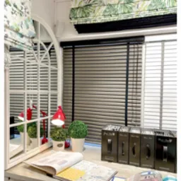 Ekspozycja różnych rodzajów rolet: rzymska w tropikalny wzór, drewniane żaluzje poziome i roleta z kwiatowym motywem, z katalogami i próbkami materiałów na blacie.