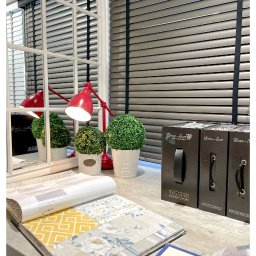 ROLETY AKCES - Ekspozycja próbek materiałów do przesłon okiennych na tle szarych żaluzji poziomych, czerwonej lampki biurkowej i dekoracyjnych roślin w doniczkach