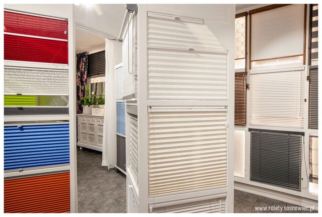 Ekspozycja różnokolorowych i różnorodnych typów osłon okiennych w salonie, widoczne plisy w odcieniach czerwieni, zieleni, niebieskiego i pomarańczowego, oraz żaluzje w kolorach białym, beżowym...