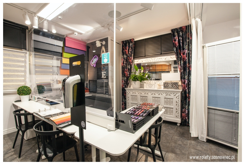 Wnętrze salonu z ekspozycją różnych typów przesłon okiennych, w tym żaluzji drewnianych i aluminiowych, z paletą kolorów na blacie i transparentną przegrodą ochronną.