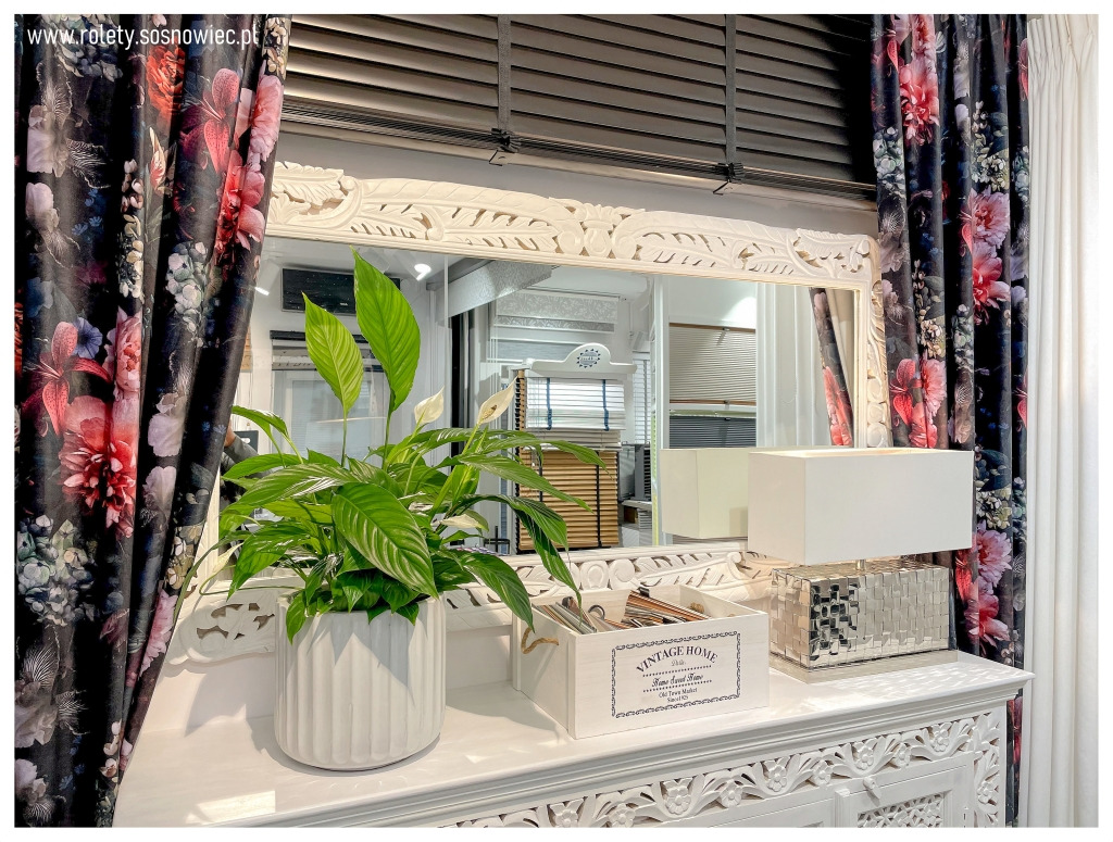 Eleganckie wnętrze z dekoracyjnymi zasłonami w kwiaty, białym lustrem w ozdobnej ramie, lampą z mozaikową podstawą i rośliną doniczkową na białej komodzie, w tle widoczne poziome żaluzje.