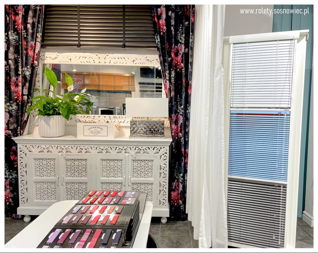 Ekspozycja różnych kolorów żaluzji aluminiowych na tle dekoracyjnej komody z lustrem i wzornikiem tkanin.