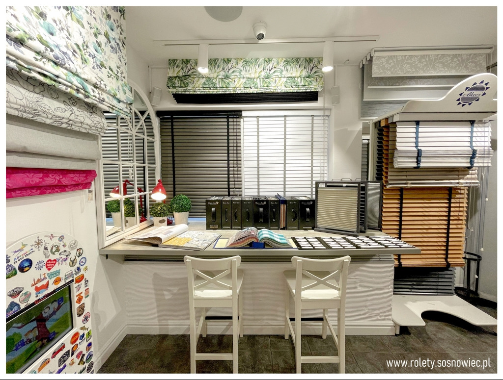 Ekspozycja różnych typów osłon okiennych w salonie w Sosnowcu:  materiały na rolety rzymskie z motywami roślinnymi, próbki żaluzji drewnianych, katalogi i próbki tkanin na blacie.