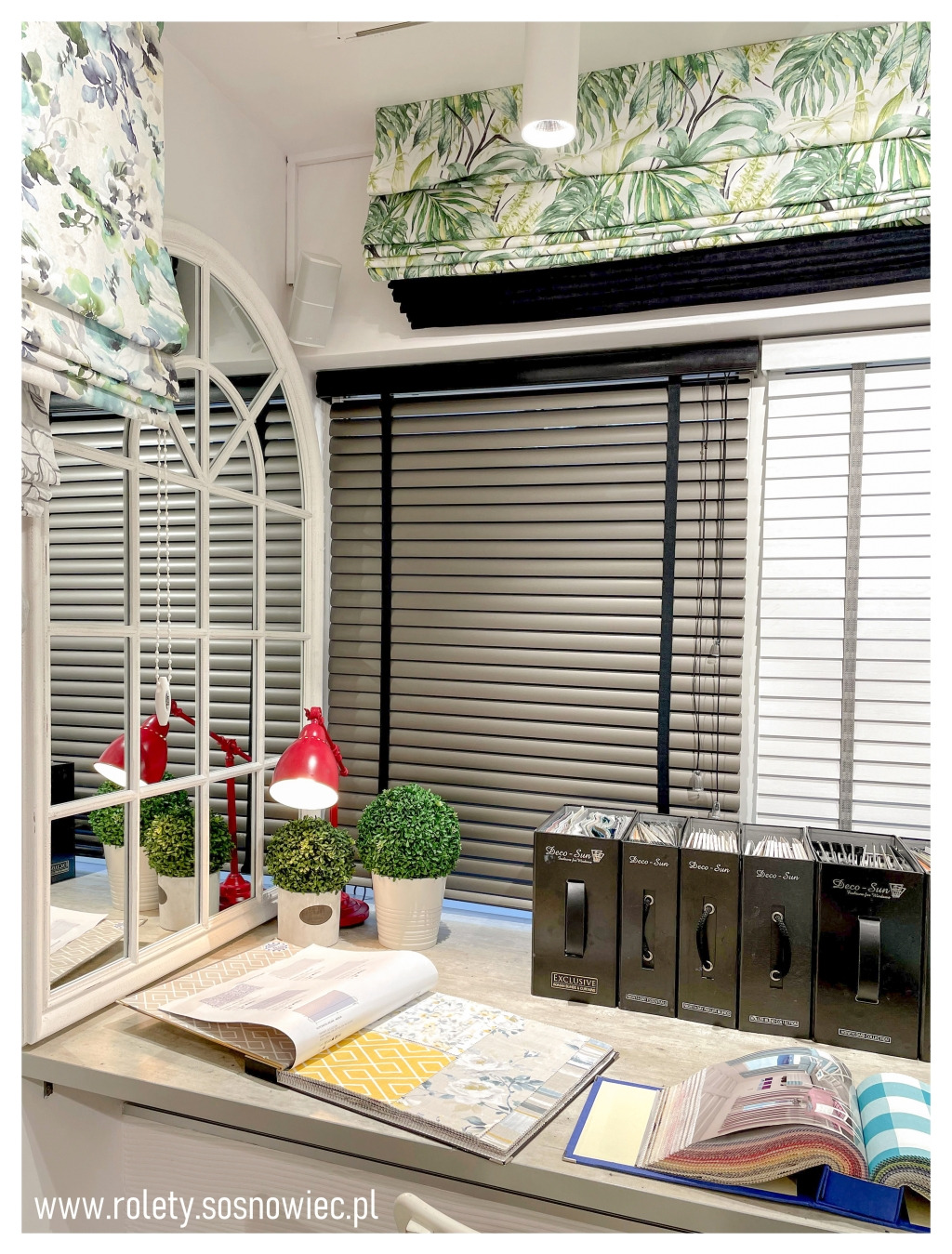 Ekspozycja różnych rodzajów rolet: rzymska w tropikalny wzór, drewniane żaluzje poziome i roleta z kwiatowym motywem, z katalogami i próbkami materiałów na blacie.