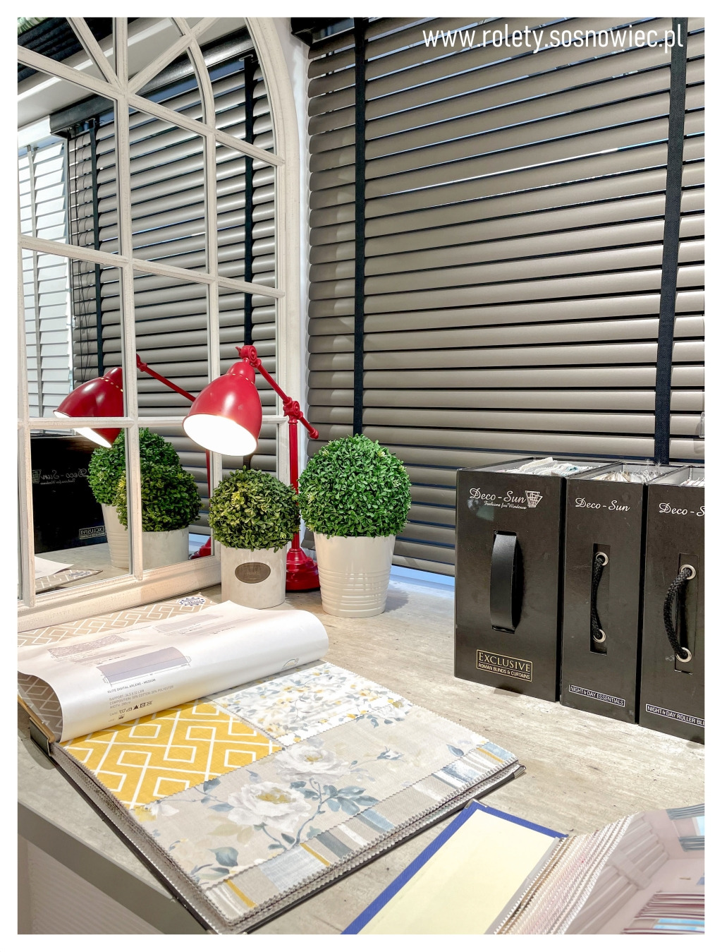 Ekspozycja próbek materiałów do przesłon okiennych na tle szarych żaluzji poziomych, czerwonej lampki biurkowej i dekoracyjnych roślin w doniczkach