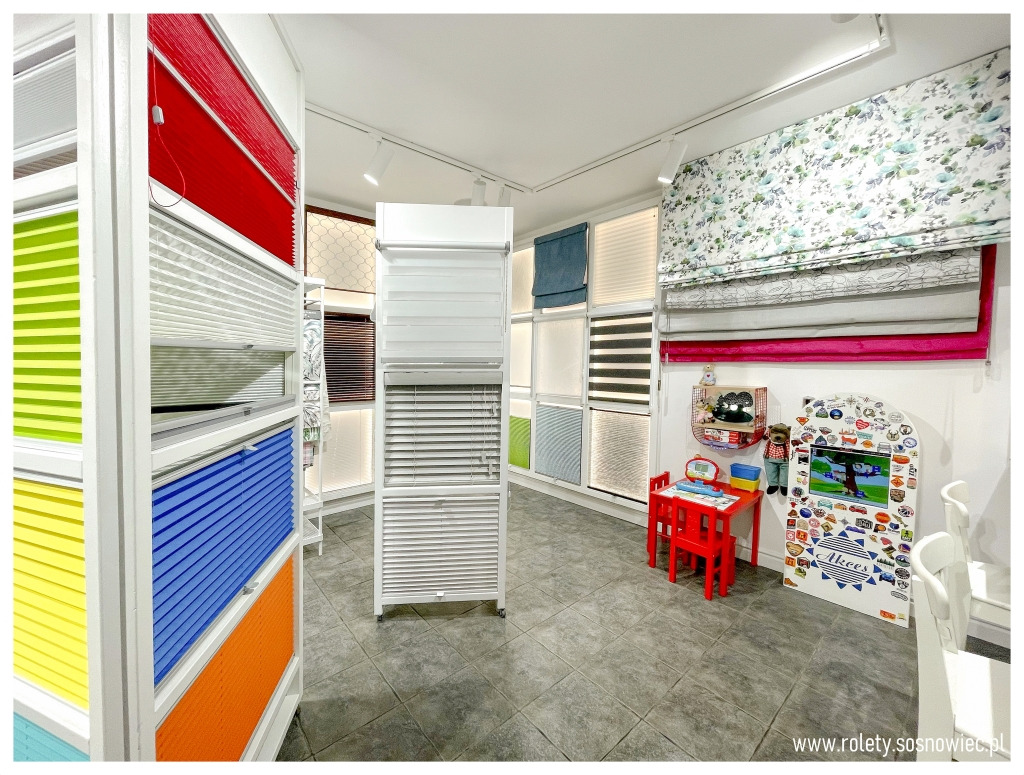 Ekspozycja różnorodnych osłon okiennych w salonie: kolorowe plisy, poziome żaluzje, tkaniny w kwiaty, aranżacja dziecięca z czerwonym stolikiem i telewizorem.