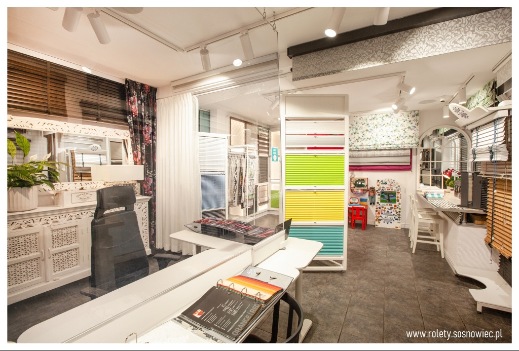 Ekspozycja różnych typów osłon okiennych w salonie: żaluzje drewniane, plisy w intensywnych kolorach, wzorniki materiałów na stole.