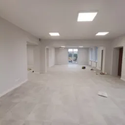 Przestronne, jasne wnętrze po remoncie, z szarymi płytkami podłogowymi i świeżo pomalowanymi na biało ścianami. Widoczne ślady po pracach wykończeniowych, takie jak resztki materiałów budowlanych...