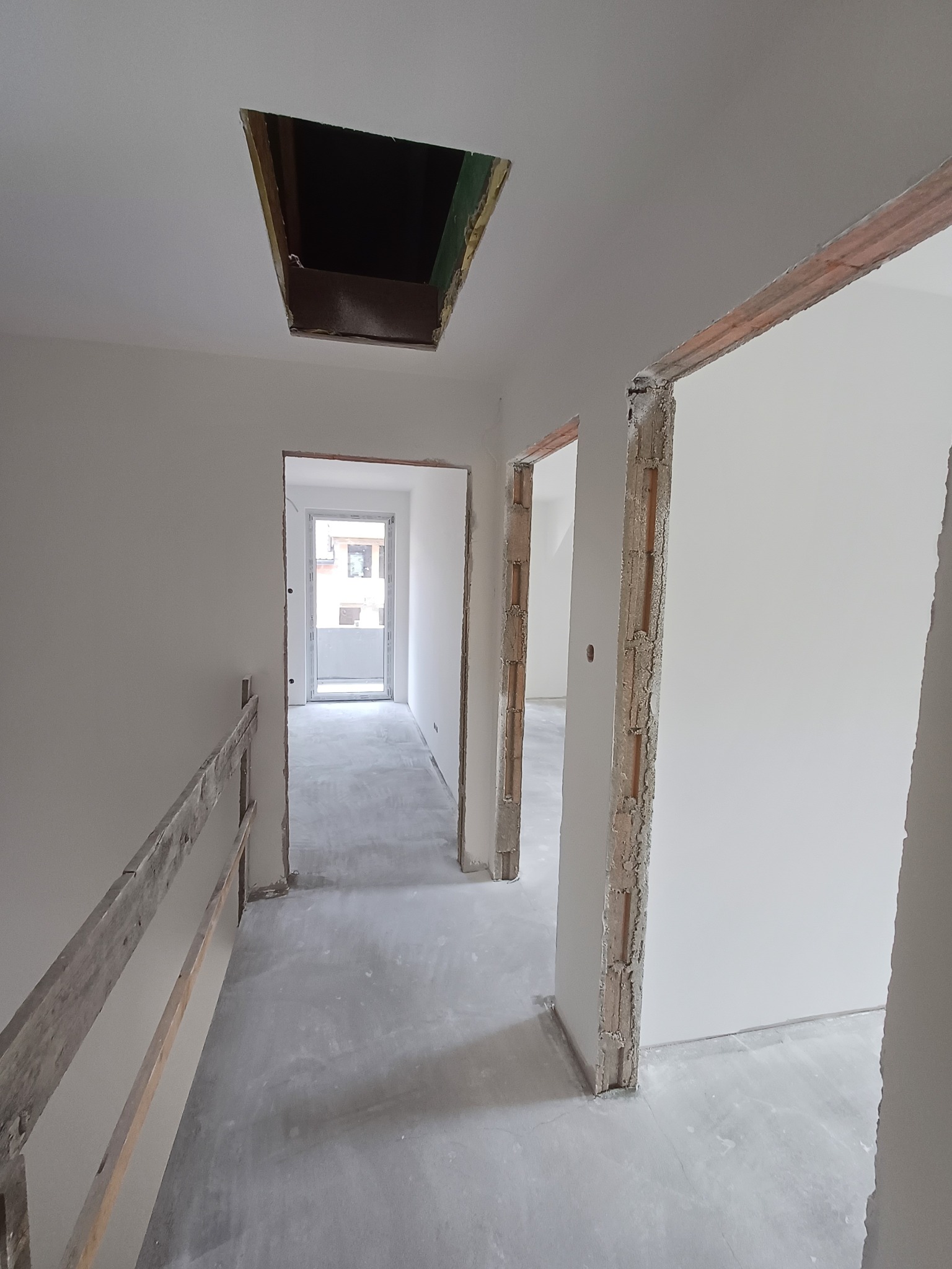Wnętrze w trakcie prac wykończeniowych: świeżo otynkowane ściany, surowy otwór w suficie, widoczne cegły w ościeżnicach i prowizoryczna drewniana barierka.