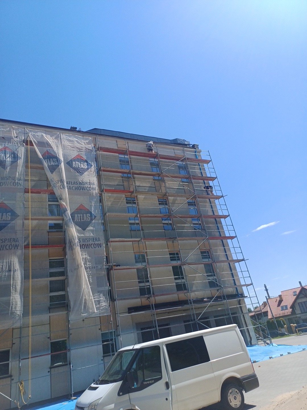 Wysoki budynek w trakcie prac elewacyjnych, osłonięty rusztowaniami i plandekami z logo Atlas, z widocznym białym busem na pierwszym planie i pracownikami na rusztowaniach.
