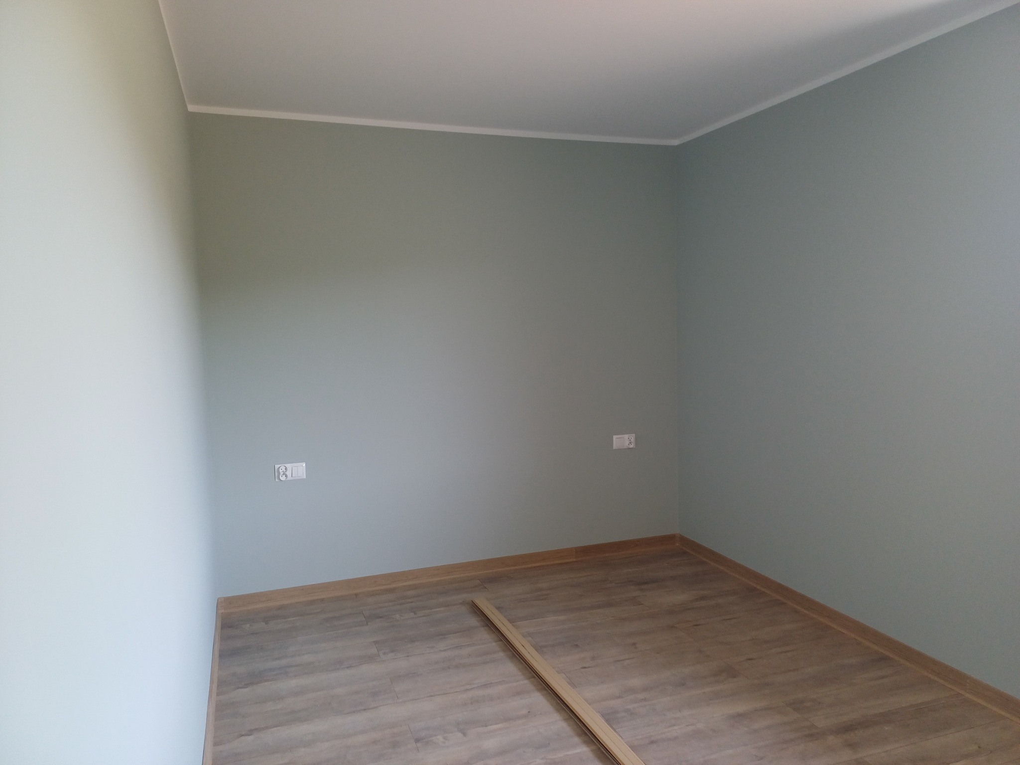 Pomalowane na szaro-zielono wnętrze pokoju z panelami podłogowymi i listwami przypodłogowymi, widoczne gniazdka elektryczne na ścianie.