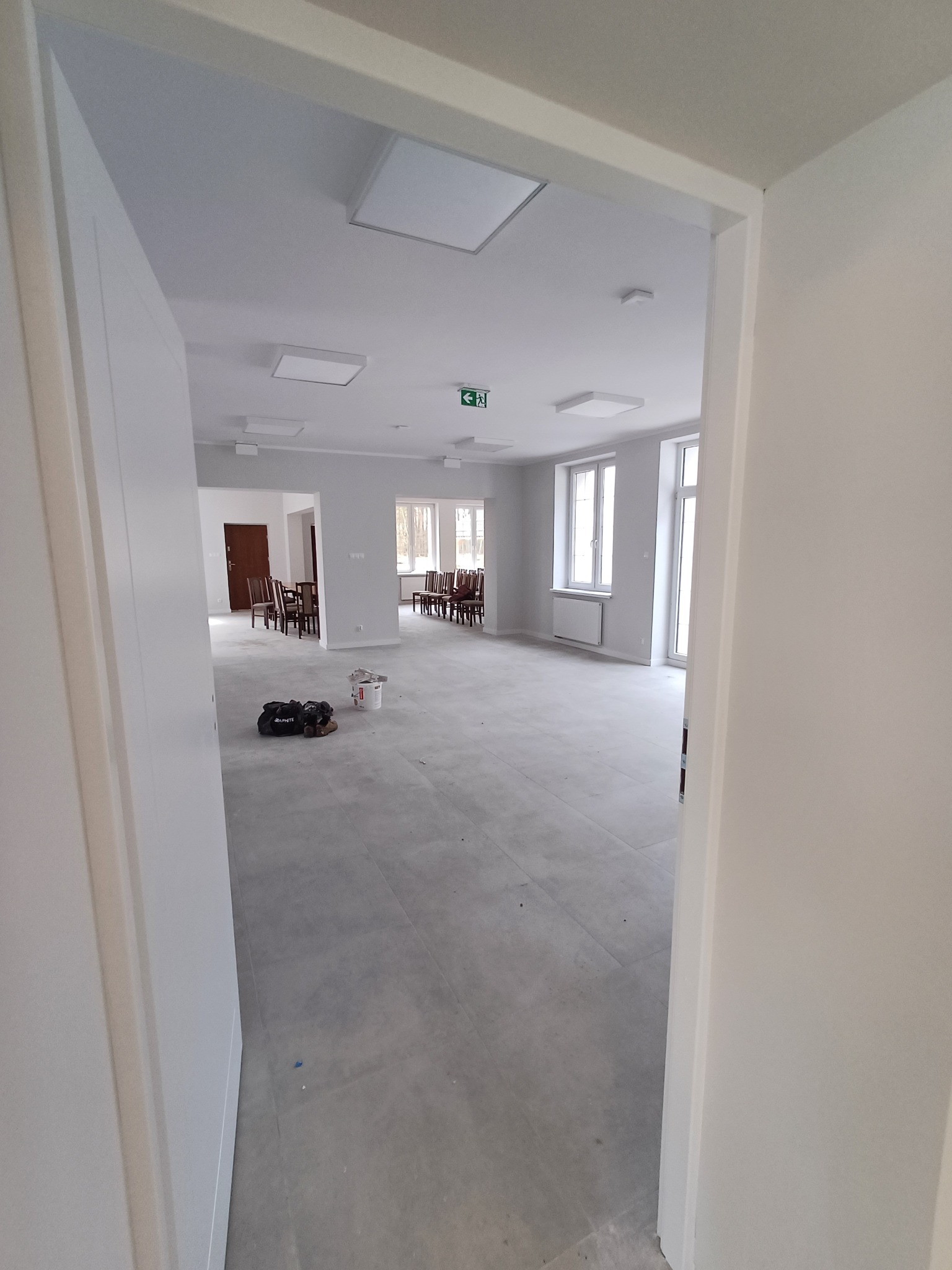 Wnętrze biura po remoncie, ściany pomalowane na biało, szary betonowy podkład, widoczne narzędzia i wiadro z farbą, okna z grzejnikami.