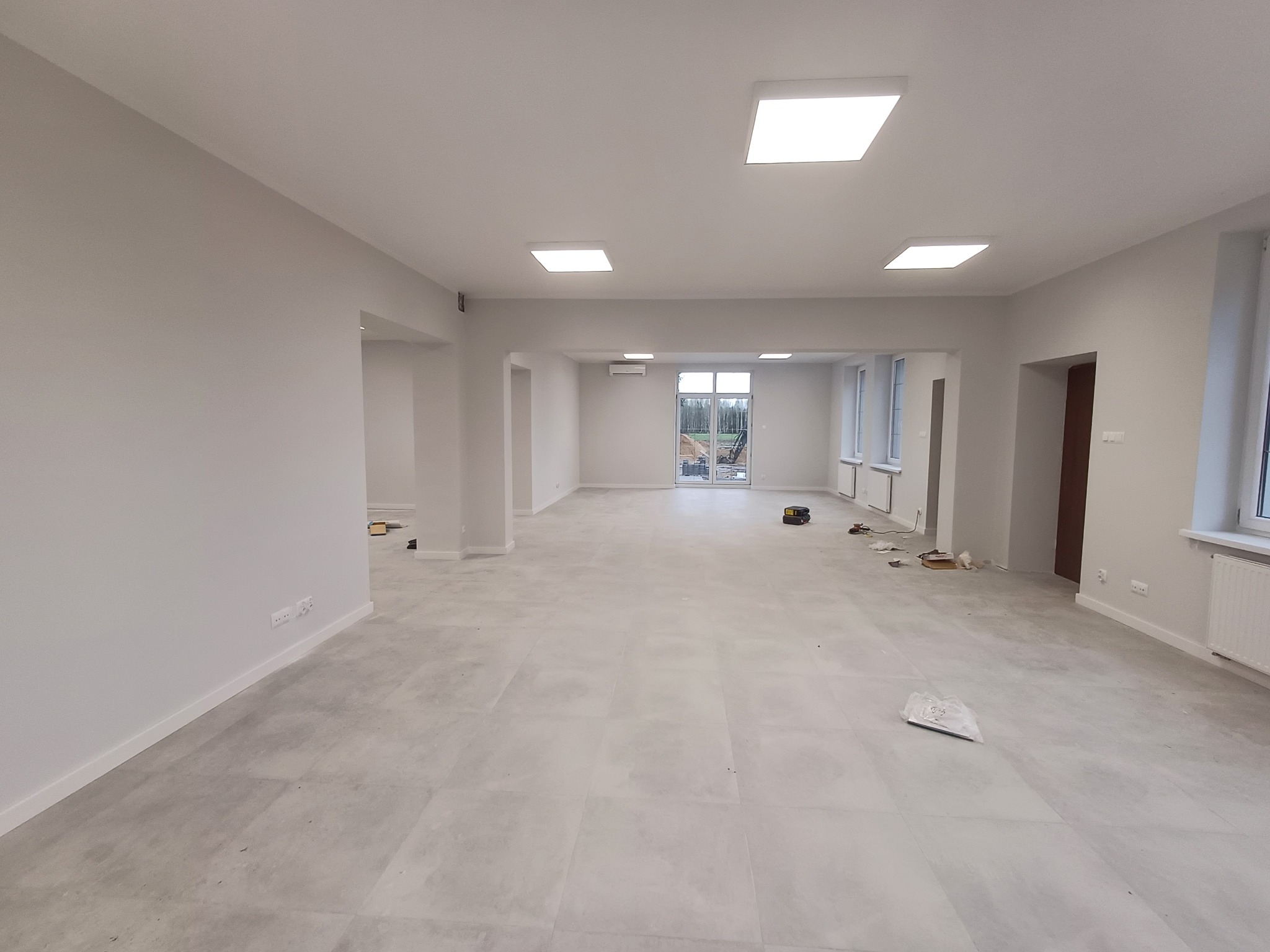 Przestronne, jasne wnętrze po remoncie, z szarymi płytkami podłogowymi i świeżo pomalowanymi na biało ścianami. Widoczne ślady po pracach wykończeniowych, takie jak resztki materiałów budowlanych...