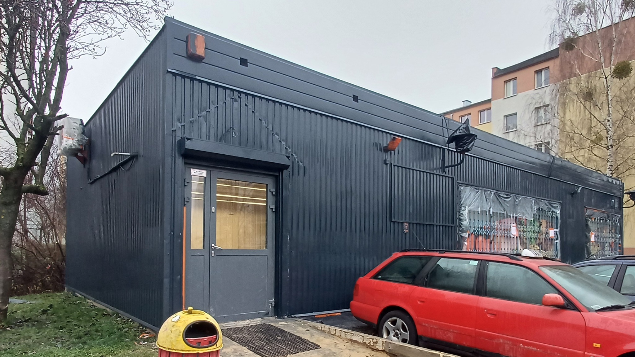 Ciemna, odnowiona elewacja budynku z blachy falistej, widoczna z zewnątrz. Czerwony samochód na pierwszym planie. Szara stolarka okienna i drzwiowa.