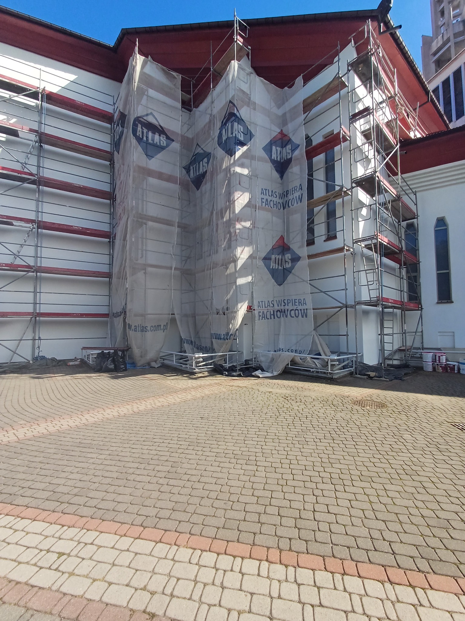 Biała elewacja budynku w trakcie prac, osłonięta siatką z logo Atlas, otoczona rusztowaniami. Widoczne puszki z farbą i kostka brukowa na pierwszym planie.