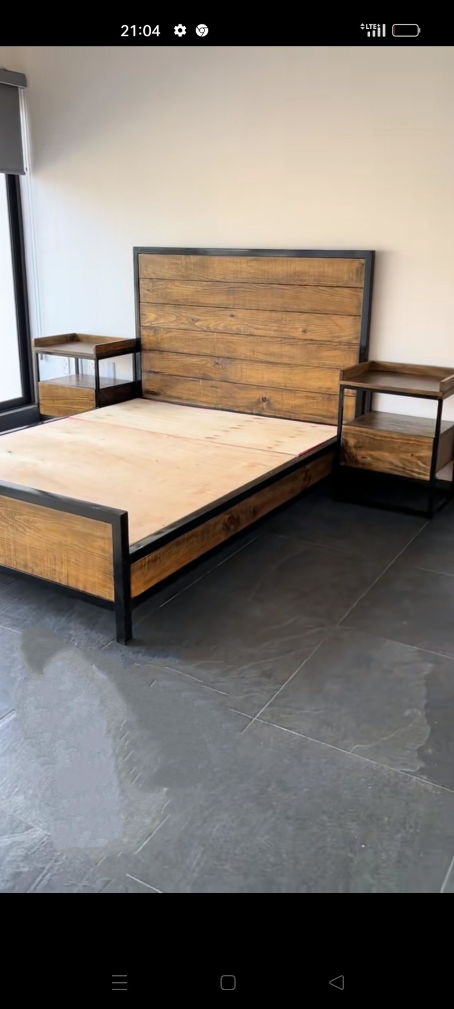 Drewniane łóżko z czarną metalową ramą i dwiema szafkami nocnymi w stylu industrialnym, na szarych płytkach podłogowych.