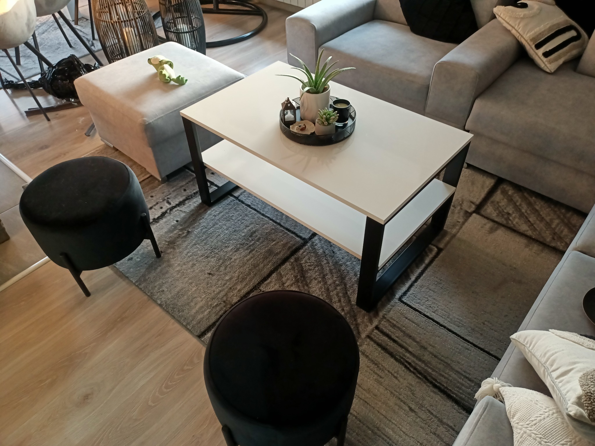 Nowoczesny salon z szarą sofą narożną, białym stolikiem kawowym z czarnymi nogami, czarnymi pufami i szarym dywanem w geometryczne wzory.