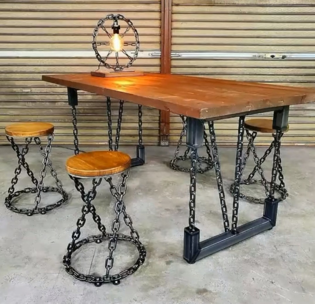 Stół z blatem z ciemnego drewna i nogami z łańcuchów oraz dopasowane hokery z siedziskami z jasnego drewna i podstawami z łańcuchów, oświetlony lampką w stylu industrialnym.