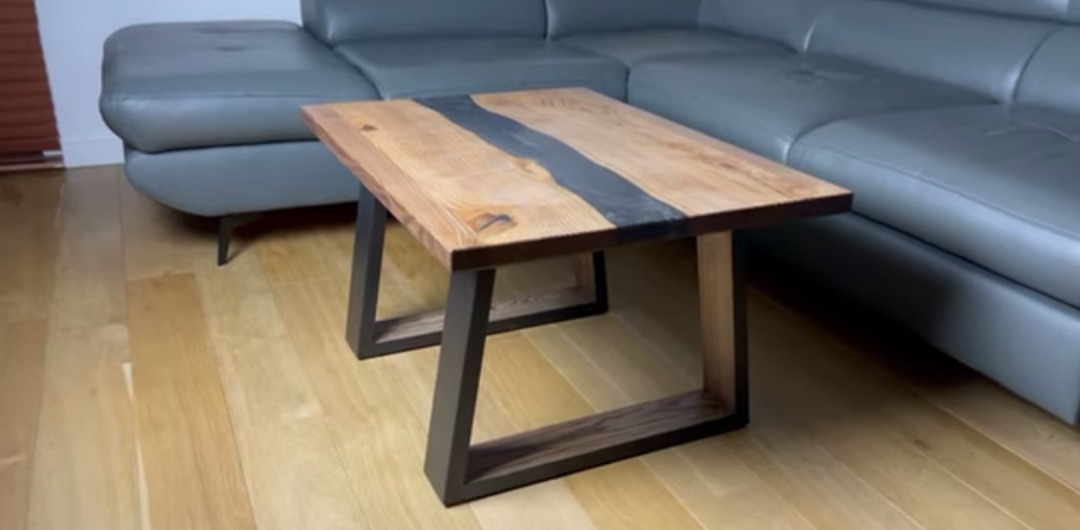 Stolik kawowy z drewnianym blatem z żywicą epoksydową w odcieniach szarości, na metalowych nogach w kształcie litery U, ustawiony na parkiecie, obok szarej sofy.