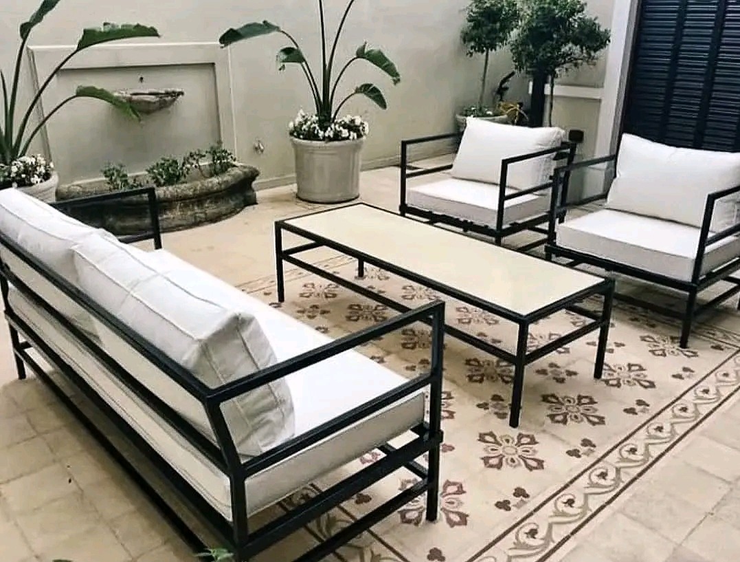 Zestaw mebli ogrodowych z czarnymi metalowymi ramami i białymi poduszkami na wzorzystej terakocie, z fontanną i roślinami w tle.