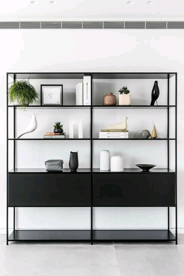 Minimalistyczna, czarna regałowa konstrukcja z szufladami, wypełniona dekoracyjnymi przedmiotami na tle białej ściany i podwieszanego sufitu.