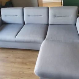 Szara, modułowa kanapa z podnóżkiem, ustawiona na drewnianej podłodze, widoczna z boku, z trzema siedziskami i subtelnymi przeszyciami.