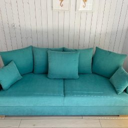 Perfect Clean Bartosz Reus - Turkusowa sofa z poduszkami na tle białej, drewnianej ściany z dekoracjami w ramkach przedstawiającymi koniki morskie.