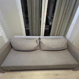 Perfect Clean Bartosz Reus - Szara, rozłożona kanapa z dwoma poduszkami na tle okna z zasłonami i ceglanej ściany z grzejnikiem.