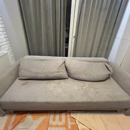 Perfect Clean Bartosz Reus - Szara kanapa z plamami na siedzisku i poduszkach, ustawiona pod oknem z zasłonami, widoczna część grzejnika i ściany z cegły.