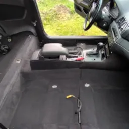 Wnętrze samochodu z wyjętymi siedzeniami, przygotowane do czyszczenia tapicerki podłogowej, widoczne pasy bezpieczeństwa, narzędzia i szczotka.