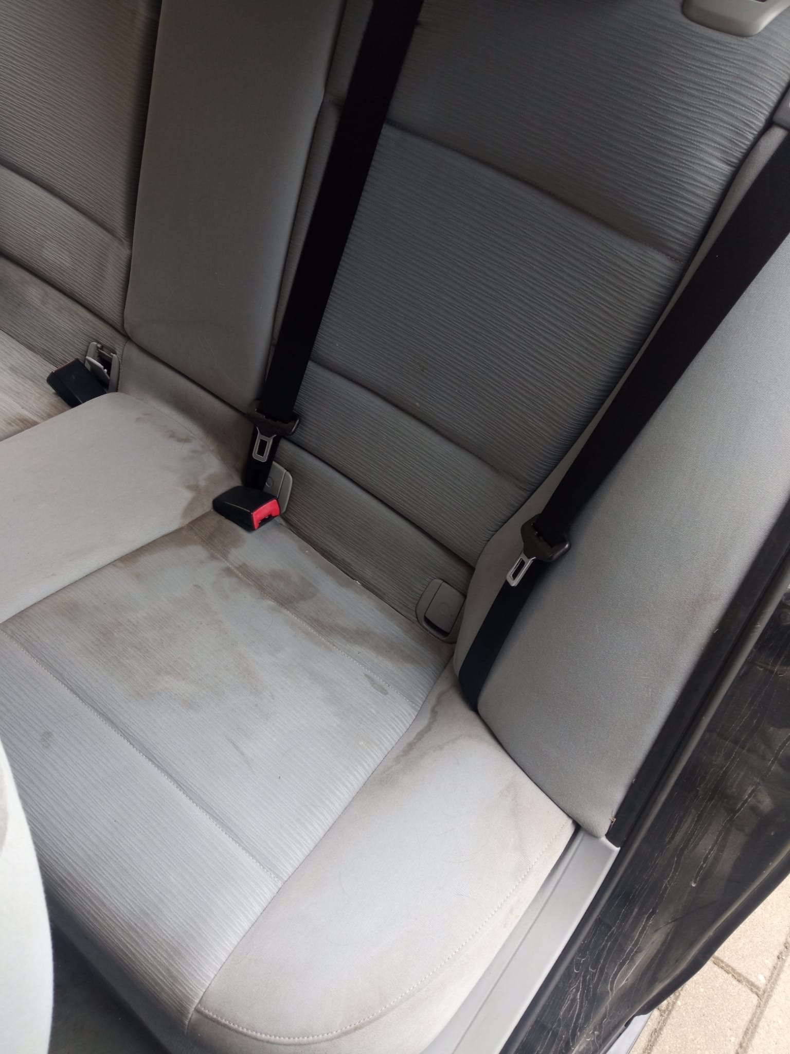 Brudna, jasnoszara tylna kanapa samochodowa z widocznymi plamami i pasami bezpieczeństwa.