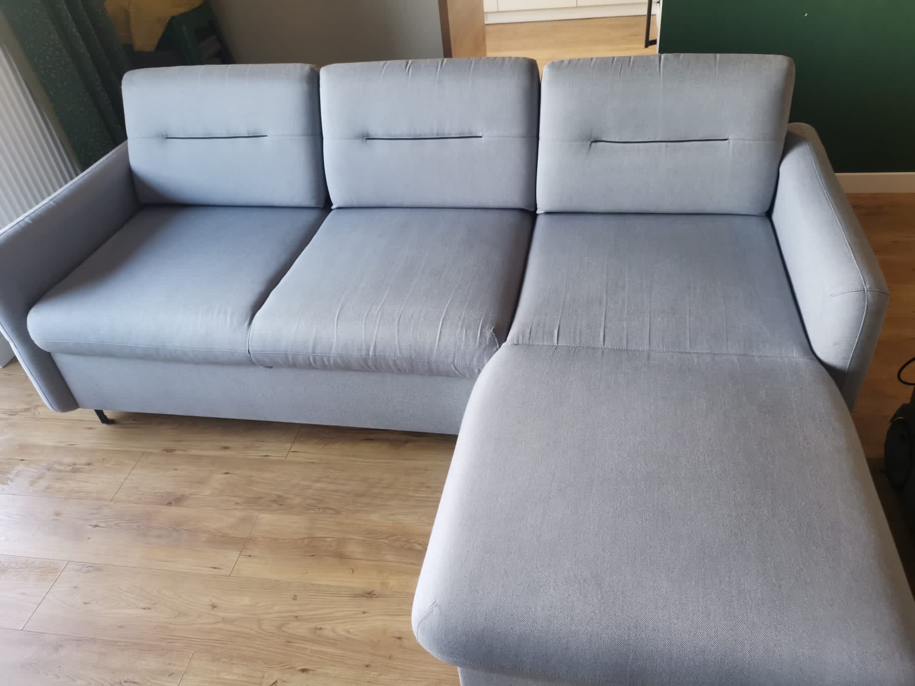 Szara, modułowa kanapa z podnóżkiem, ustawiona na drewnianej podłodze, widoczna z boku, z trzema siedziskami i subtelnymi przeszyciami.